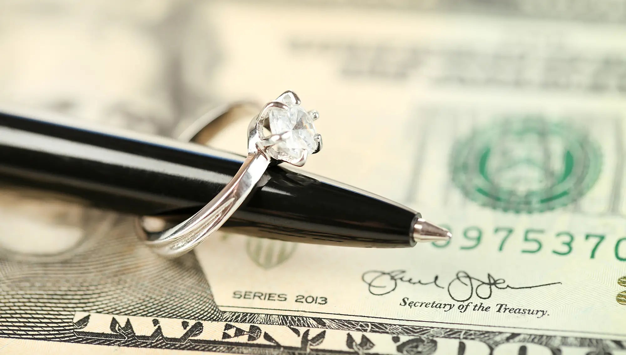 Diamond ring on black pen over money, symbolizing layaway and financing options at Joe Kassab Jewelers
